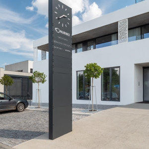 Venkovní orientační totem s hodinami, logem obce a seznamem institucí, instalovaný u vstupu do budovy